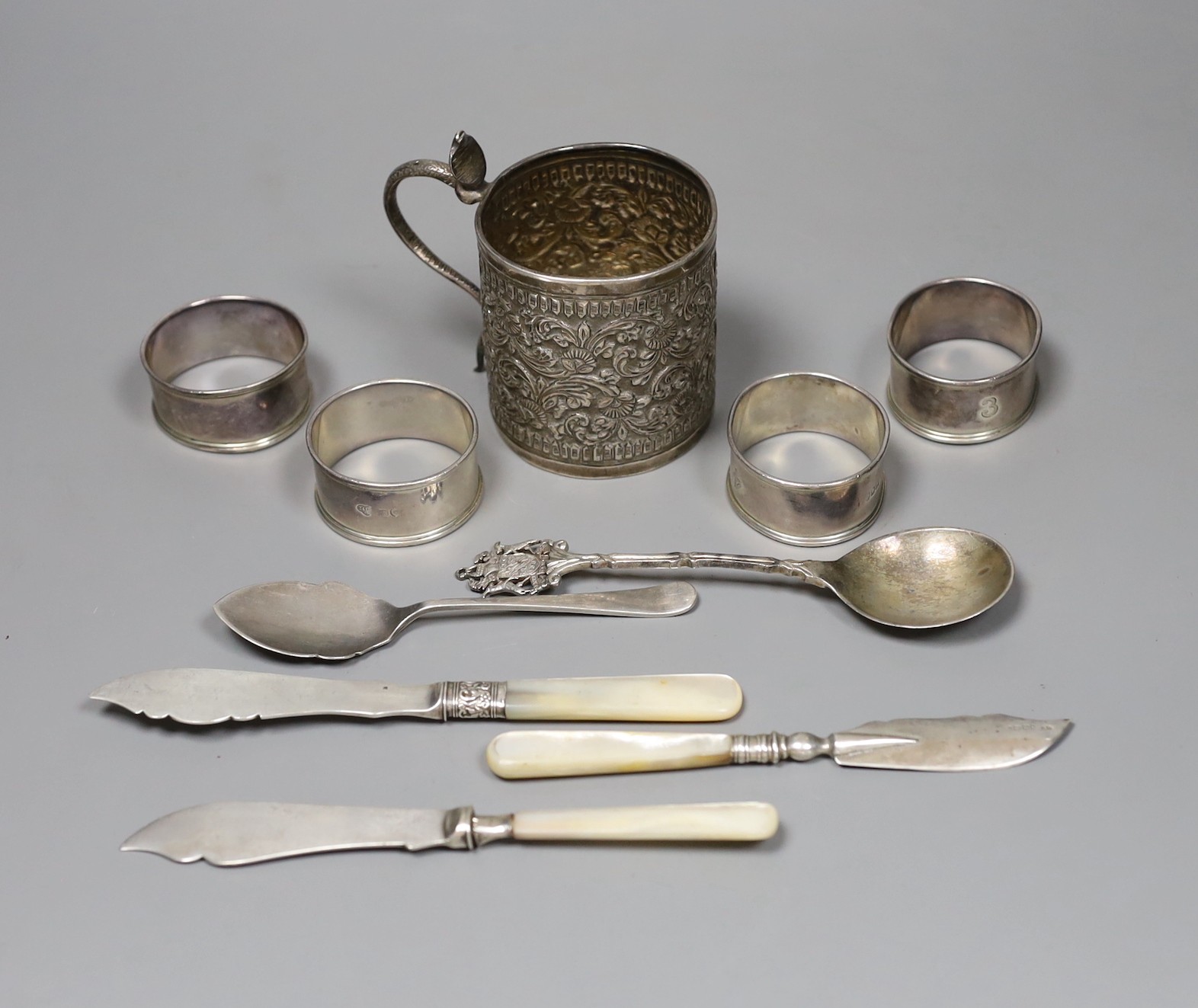 A 19th century Indian white metal christening can with cobra handle, a set of four silver napkin rings, two spoons and three mother of pearl handled butter knives.